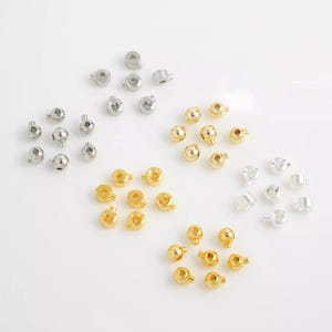 Puede incluir: Un surtido de pequeñas cuentas metálicas en tonos dorados y plateados. Las cuentas son redondas con un pequeño agujero para ensartar. Las cuentas están dispersas sobre una superficie blanca, ofreciendo una variedad de opciones para la fabricación de joyas o proyectos de artesanía.