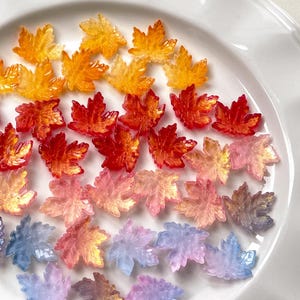 Puede incluir: Una colección de adornos en forma de hojas de arce translúcidas y coloridas. Las hojas están dispuestas sobre un plato blanco, mostrando un degradado de colores que va del naranja al rojo, rosa, azul y morado. Ideales para proyectos de manualidades.