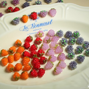 Puede incluir: Una colección de coloridos abalorios de frutas, que incluyen frambuesas naranjas, fresas rojas, uvas moradas y moras rosas, están dispuestas sobre un plato blanco. El plato tiene texto azul que dice "Le Bonmasel".