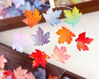 May include: A collection of colorful maple leaf charms in various shades of orange, red, purple, blue, and green. The translucent leaves are arranged on a clear surface, with a wooden frame in the background. These charms are ideal for jewelry making or craft projects.