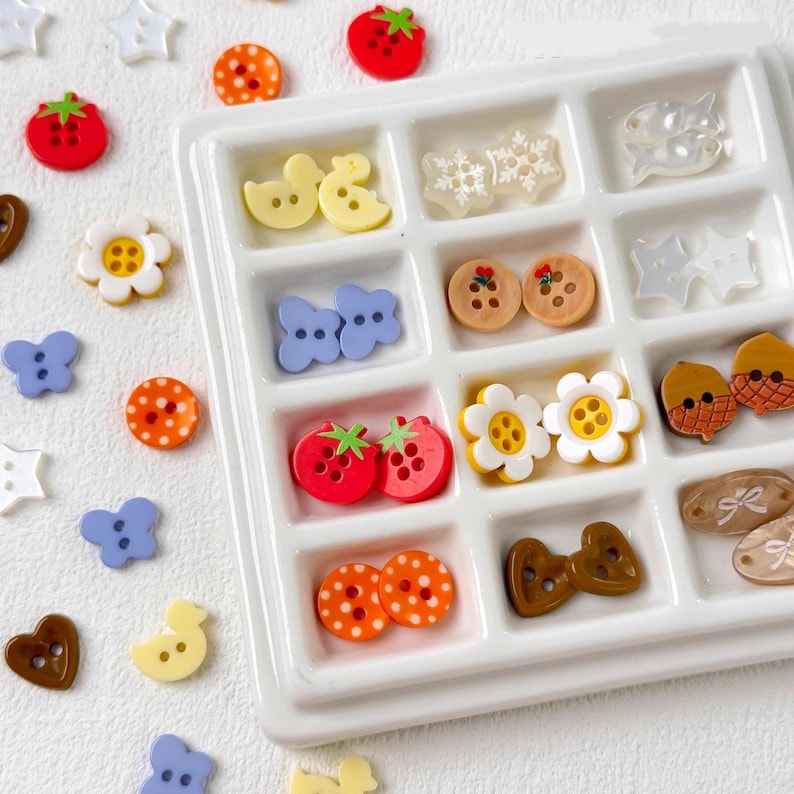 May include: A collection of colorful decorative buttons in various shapes, including flowers, butterflies, and fruits. The buttons are arranged in a white tray and scattered around it. The buttons are in shades of red, orange, yellow, blue, and brown.