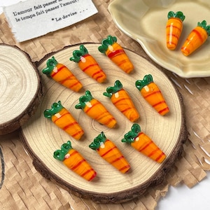 Puede incluir: Un conjunto de cuentas de vidrio en miniatura con forma de zanahoria. Cada cuenta presenta un cuerpo naranja con rayas rojas y una parte superior verde con hojas. Las cuentas están dispuestas sobre una rodaja de madera y un plato pequeño.