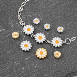 Op de afbeelding: Een zilveren ketting met madeliefjesbedels en verschillende losse madeliefjesbedels. De madeliefjesbedels hebben witte bloemblaadjes, grijze omtrekken en oranje centra. De ketting en bedels liggen op een donkergrijze ondergrond.