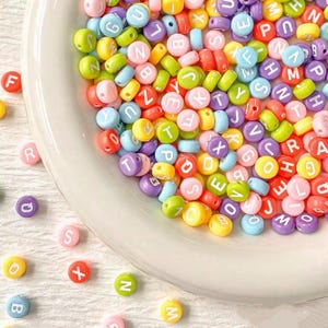 May include: A selection of colourful alphabet beads in a white bowl and scattered on a white surface. The round beads feature letters in various colours, including pink, yellow, green, and purple. Suitable for jewellery making and crafts.
