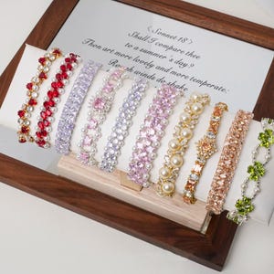 Può includere: Una selezione di braccialetti gioiello in vari design e colori, tra cui rosso, viola e rosa. Alcuni braccialetti presentano perle e pietre preziose. I braccialetti sono esposti su una superficie bianca all'interno di una cornice di legno, con una poesia stampata sullo sfondo.