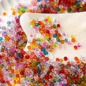 以下が含まれることがあります： 赤、オレンジ、黄色、緑、青、紫、ピンクの色合いをした、小さく半透明の多色ガラスシードビーズの大きなコレクション。ビーズは白い表面に散らばっており、一部は湾曲した白いトレイの上に置かれています。