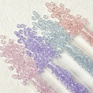 May include: Close-up of four clear plastic tubes filled with colorful beads. The beads are in shades of pink, light blue, and purple. The beads are scattered on a white textured surface. Ideal for jewelry making and crafting.