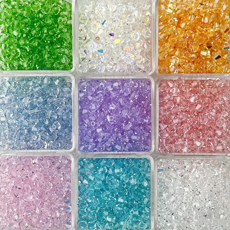 May include: A close-up view of a nine-compartment container filled with colorful crystal beads. The colors include green, clear, orange, blue, purple, pink, light pink, turquoise, and white. The beads are small and faceted, reflecting light.