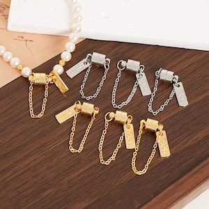 Peut inclure: Plusieurs fermoirs magnétiques pour bijoux, dorés et argentés. Chaque fermoir est orné d'une étiquette rectangulaire gravée, reliée à une chaîne. Les fermoirs sont disposés sur une surface en bois, avec un collier de perles visible.