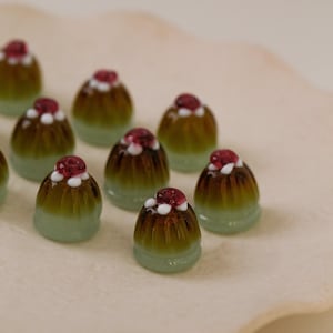 May include: A collection of miniature glass desserts. Each dessert features a light green base, a dark brown center, and a red and white topping. The desserts are arranged on a light-colored surface.