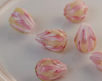 May include: Close-up of several glass beads with a floral design. The beads are a mix of pink, white, and yellow, with a translucent quality. The beads are arranged on a white surface. These beads are suitable for jewelry making.