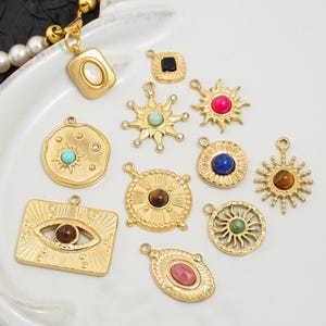 Pode incluir: Uma coleção de pingentes dourados com vários designs. Os pingentes apresentam pedras preciosas em cores como turquesa, rosa e marrom. Alguns pingentes têm desenhos de sol e olho. Os pingentes estão em uma superfície branca.