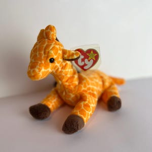 May include: A plush giraffe toy with an orange and white patterned body, brown hooves, and black eyes. The toy has a tag attached with a red heart and the words "Beanie Baby".