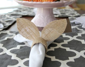 May include: A burlap napkin ring shaped like bunny ears, with a white napkin tucked inside. The ring is on a grey and white patterned table runner.