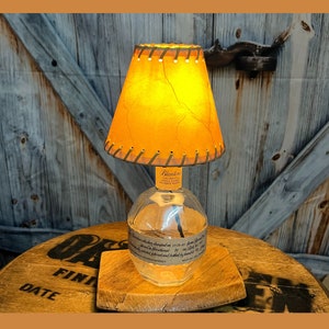 May include: A repurposed whiskey bottle lamp with a brown leather lampshade. The lamp is sitting on a wooden base with the words "Date" and "Finish" visible. The lamp is lit and casting a warm glow.