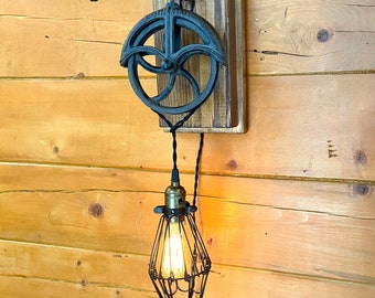 May include: A rustic industrial style light fixture with a black metal pulley and cage light bulb hanging from a wooden wall mount.