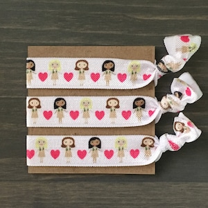 May include: Four white elastic hair ties with a pattern of girls and pink hearts. The hair ties are on a brown card.