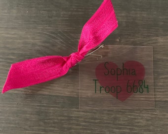 May include: A clear plastic tag with a pink heart and the text "Sophia Troop 6684". A pink ribbon is attached to the tag.