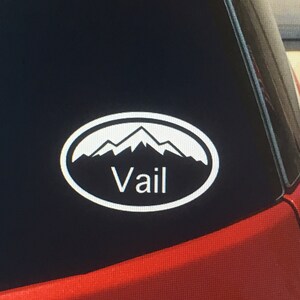May include: White oval sticker with a mountain range silhouette and the word "Vail" in white text on a black car window.