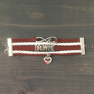 May include: A brown and white braided bracelet with a silver charm that says "Love Brownie". The charm is attached to a silver infinity symbol. The bracelet has a silver heart charm hanging from it.