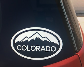May include: White oval sticker with a mountain range silhouette and the word "COLORADO" in white text on a black car window.