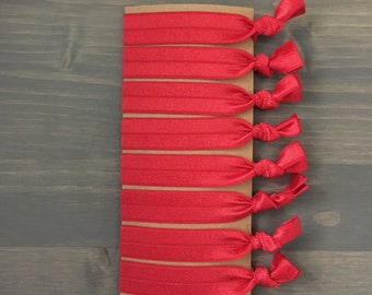 May include: A set of eight red hair ties, packaged on a brown card. The hair ties are made of a stretchy material and have a knotted design.