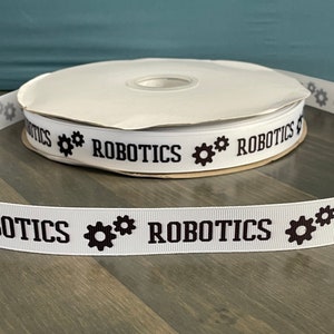 May include: White ribbon with black text that reads "Robotics" and a black gear symbol on either side of the text. The ribbon is rolled up on a white spool.