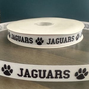 May include: A roll of white ribbon with the word "JAGUARS" printed in black, along with black paw print graphics. The ribbon is displayed on a reflective surface.