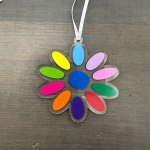May include: A colorful, clear plastic ornament shaped like a flower with a white ribbon for hanging. The flower has ten petals, each a different color: yellow, green, pink, orange, purple, red, blue, light blue, lavender, and dark blue.