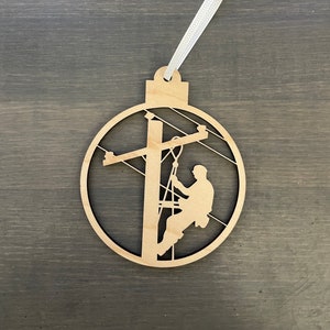 May include: A wooden ornament shaped like a circle with a silhouette of a person climbing a utility pole with power lines. The ornament has a white ribbon attached to the top.