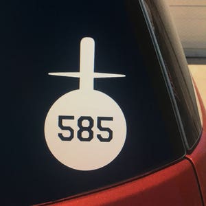 May include: White vinyl sticker with a cross and a circle containing the number 585. The sticker is on a red car window.