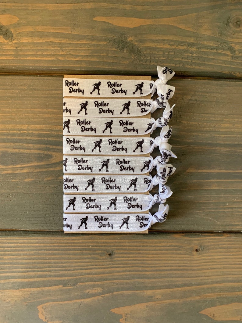 May include: A set of white elastic hair ties with black roller derby skater silhouettes and the text "Roller Derby" repeated on each tie.