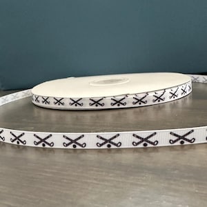 May include: White ribbon featuring a repeating pattern of black golf clubs and balls. The ribbon is rolled on a spool, resting on a wooden surface. The background is a teal colour.