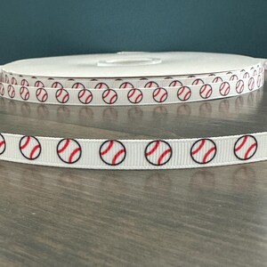 May include: White ribbon with a repeating pattern of red and black baseballs. The ribbon is spooled and also shown unraveled, displaying the baseball design. The ribbon is on a wooden surface.