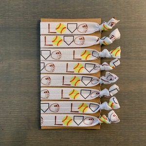 May include: Seven white elastic hair ties with a softball design. Each tie displays the word "LOVE", a softball, a baseball diamond, and a baseball glove. The ties are attached to a brown cardboard backing.
