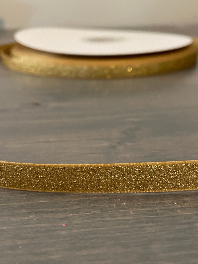 May include: A close-up of gold glitter ribbon. The ribbon is rolled up and has a white label.