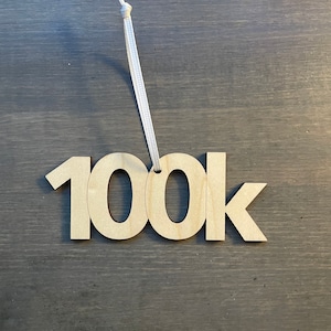 May include: A wooden ornament spelling out "100k" with a white ribbon for hanging. The numbers and letter are cut from light-colored wood and are attached with a white ribbon. The ornament is displayed on a dark gray surface.