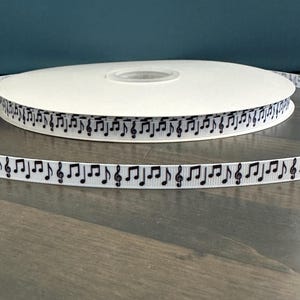 May include: A roll of white ribbon featuring a repeating pattern of black musical notes and treble clefs. The ribbon is displayed on a wooden surface, with a dark teal background.