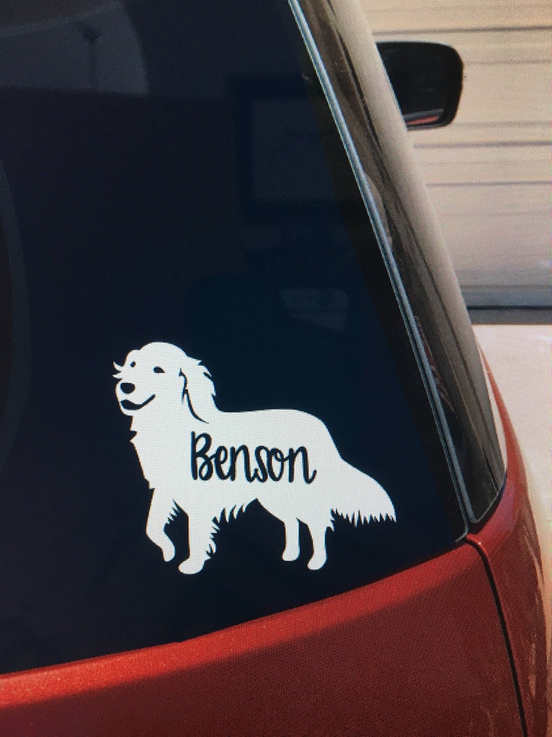 May include: A white vinyl decal of a Golden Retriever dog with the name "Benson" in a script font. The decal is applied to the rear window of a red vehicle. The dog is depicted in a walking pose.