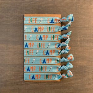 May include: A set of eight blue elastic hair ties with a white and orange print of a tent, a campfire, and the words "Camp Life".