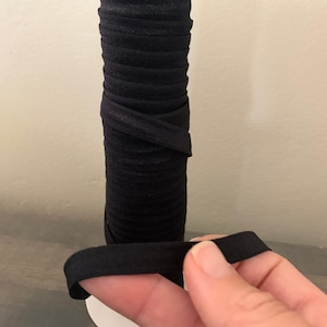 May include: A spool of black elastic fabric, wound tightly and held by a white stand. A hand is holding a piece of the elastic fabric.