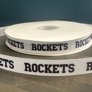 May include: White ribbon spool with the word "ROCKETS" printed in black. The ribbon is displayed on a wooden surface with a dark teal background. The ribbon is likely used for crafting, gift wrapping, or sports-themed projects.
