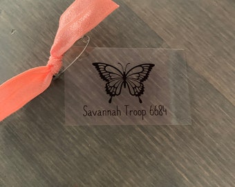 May include: A clear plastic tag with a black butterfly design and the text "Savannah Troop 6684". A pink elastic band is attached to the tag with a safety pin.