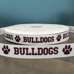 May include: White ribbon with the word "BULLDOGS" printed in maroon with paw prints on either side. The ribbon is rolled up on a spool.