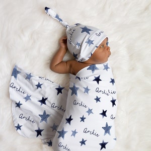 May include: A baby swaddled in a white and blue star patterned swaddle with the name "Archie" printed on it. The baby is wearing a matching white and blue star patterned knotted hat with the name "Archie" printed on it.