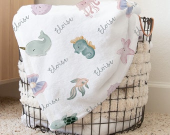 May include: White baby blanket with a seahorse, starfish, turtle, jellyfish, narwhal, and fish design. The blanket has the name "Eloise" written on it multiple times.