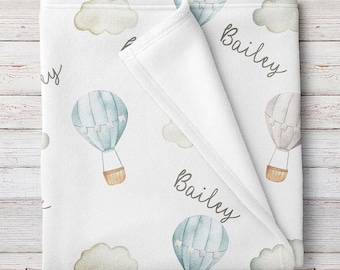 May include: A white baby blanket with a watercolor design of hot air balloons and clouds. The blanket is personalized with the name "Bailey" written in cursive.