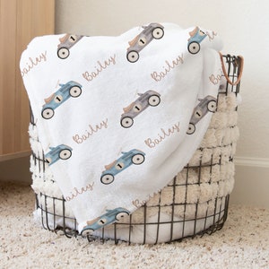 Peut inclure: Une couverture de bébé blanche avec un motif répété de voitures jouets bleues et grises. La couverture porte le nom "Bailey" imprimé en cursive brune.