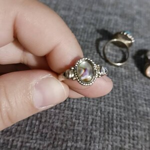 May include: A silver ring with an oval abalone shell center stone. The ring has a beaded border and decorative accents on the sides. Other rings are visible in the background.