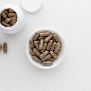Puede incluir: Un tazón blanco lleno de cápsulas marrones. El tazón está sobre una superficie blanca. Hay una botella blanca con tapa y más cápsulas marrones en el fondo.